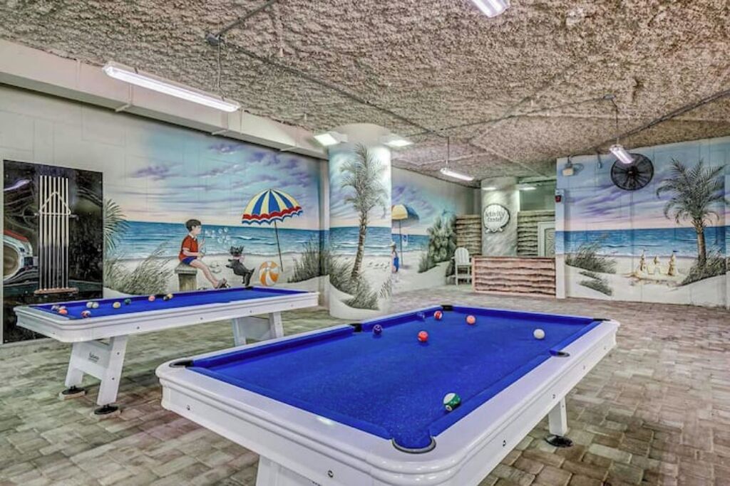 Un espace piscine comprenant deux tables de billard, une fresque représentant une scène de plage et un ventilateur de plafond.