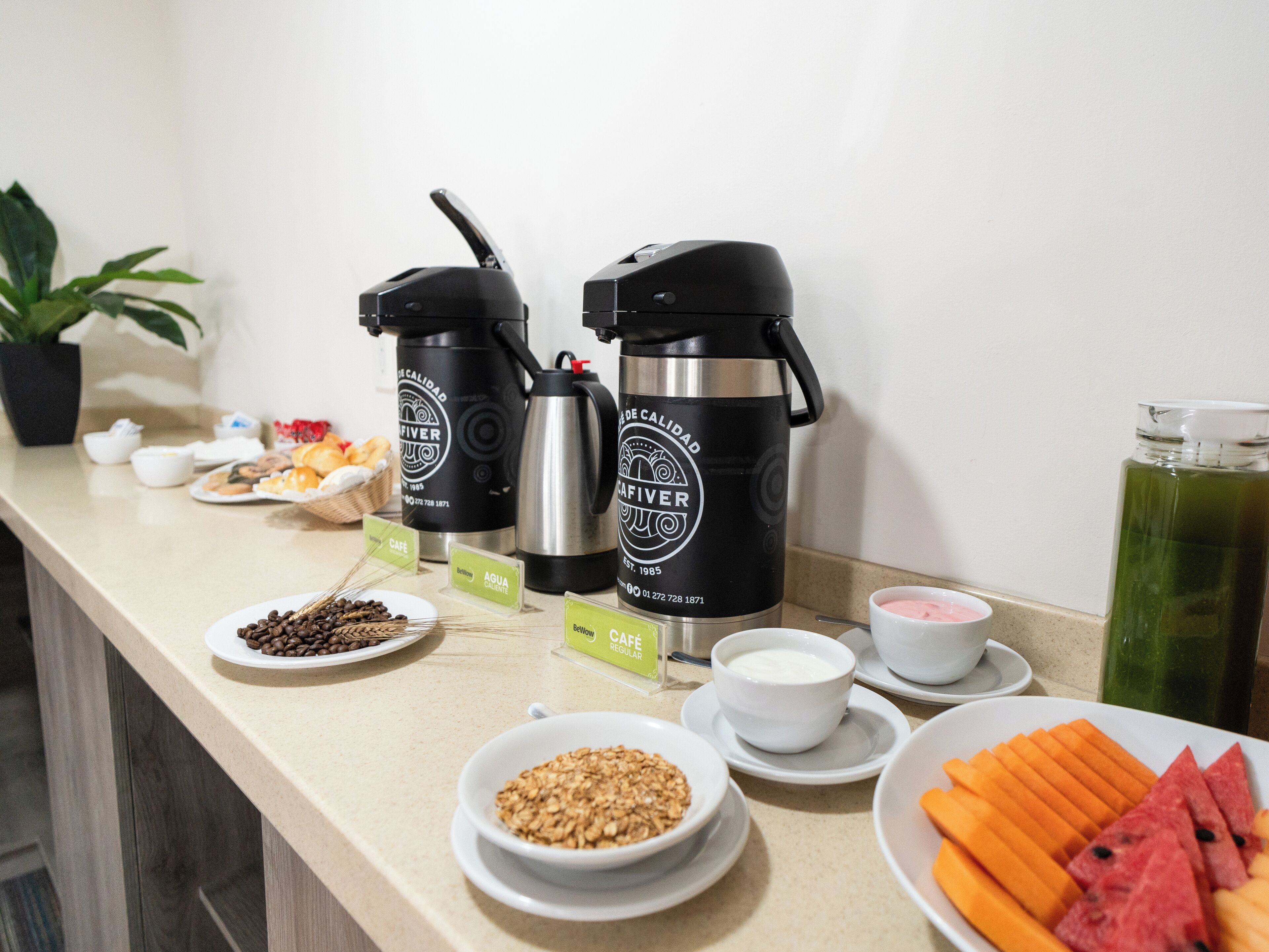 Una mesa de buffet con cafeteras, fuentes de comida y una planta.