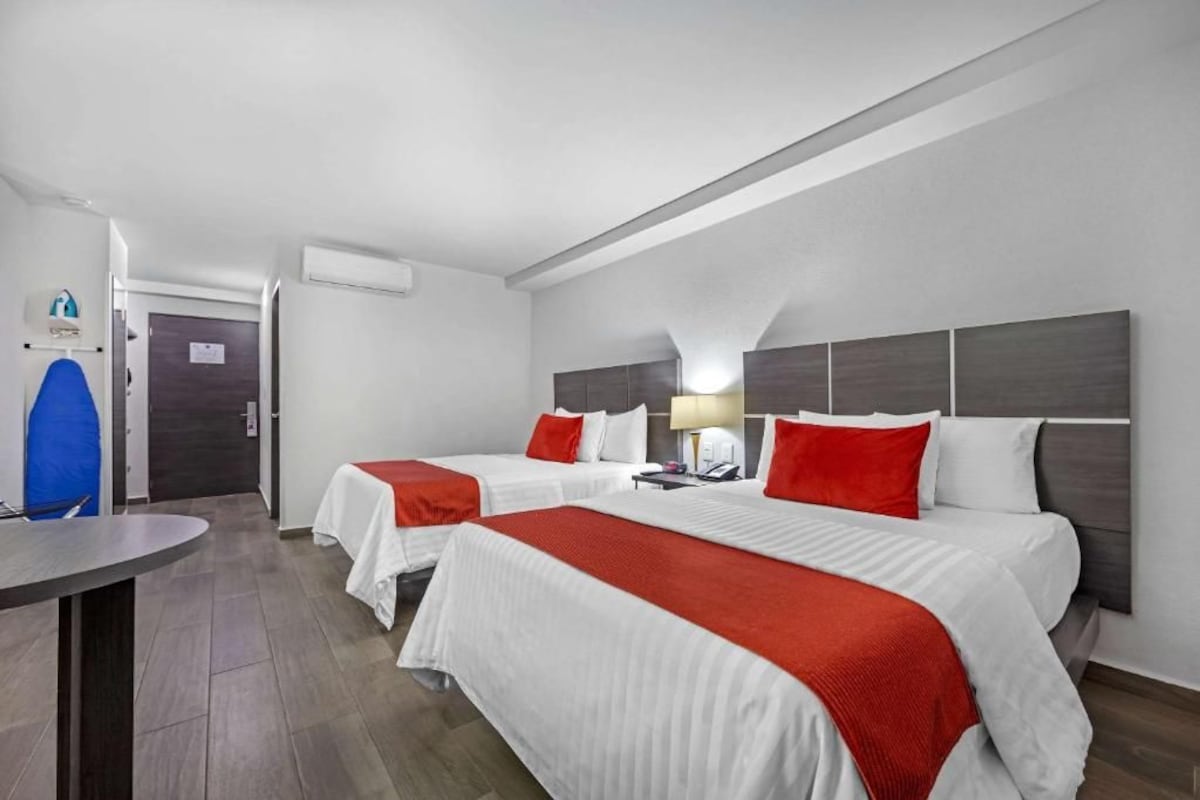 A hotel room with two beds, each with white bedding and red accents, a bedside table with a lamp, and a small desk with a chair.