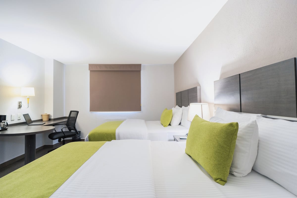 A hotel room with two beds, a desk with a chair, a lamp, and a window with a blind.