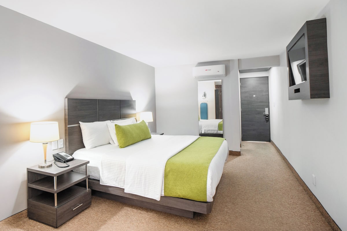 A modern hotel room with a bed, bedside table, lamp, and a flat-screen TV mounted on the wall.