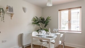Ein Essbereich mit einem weißen Tisch, gedeckt für vier Personen, einer Zimmerpflanze und einem Fenster mit Jalousien.