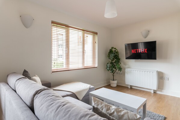 Un moderno salón con un sofá gris, una mesa de centro blanca, una planta en maceta, un radiador y un televisor montado en la pared mostrando 'NETFLIX'.