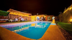 Ein Schwimmbeckenbereich bei Nacht mit überdachter Sitzgelegenheit und einem Gebäude im Hintergrund.