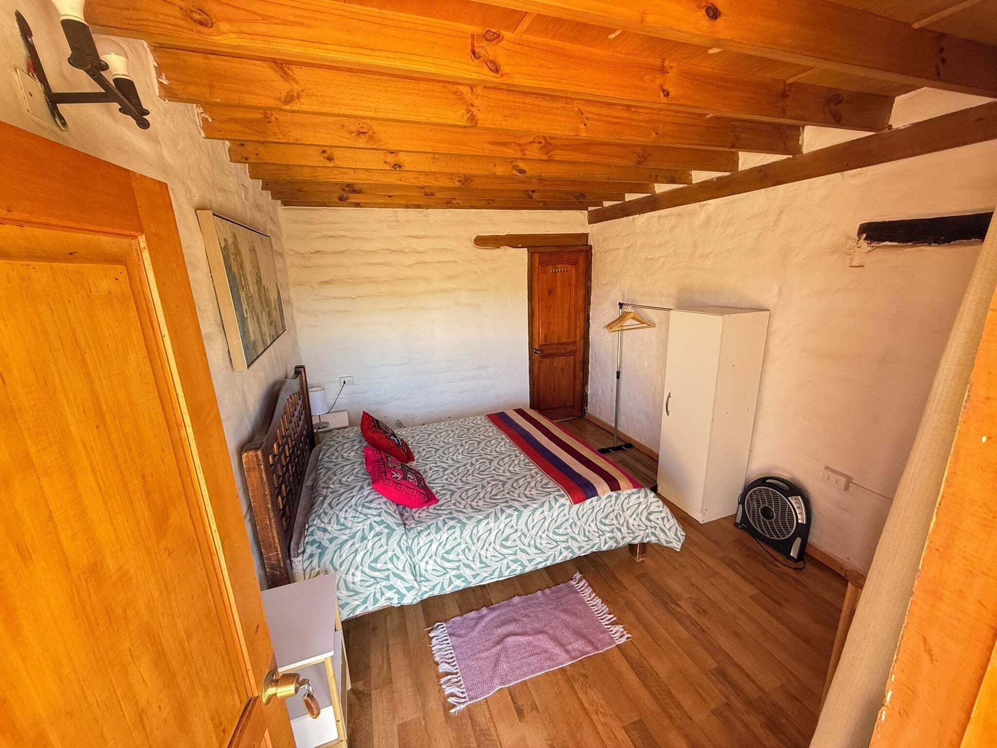 Una habitación acogedora con techo de madera, una cama con ropa de cama estampada, un armario de madera y un pequeño ventilador en el suelo.
