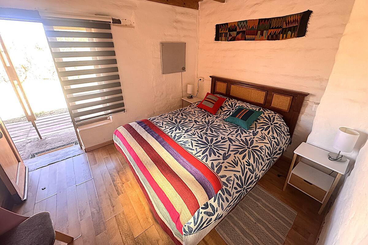 A bedroom with a bed, a bedside table, a nightstand, a window with blinds, and a wall-mounted artwork.
