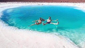 Zwei Personen treiben in einem großen, klaren blauen Pool, umgeben von weißem Sand, mit einer kargen Landschaft im Hintergrund.