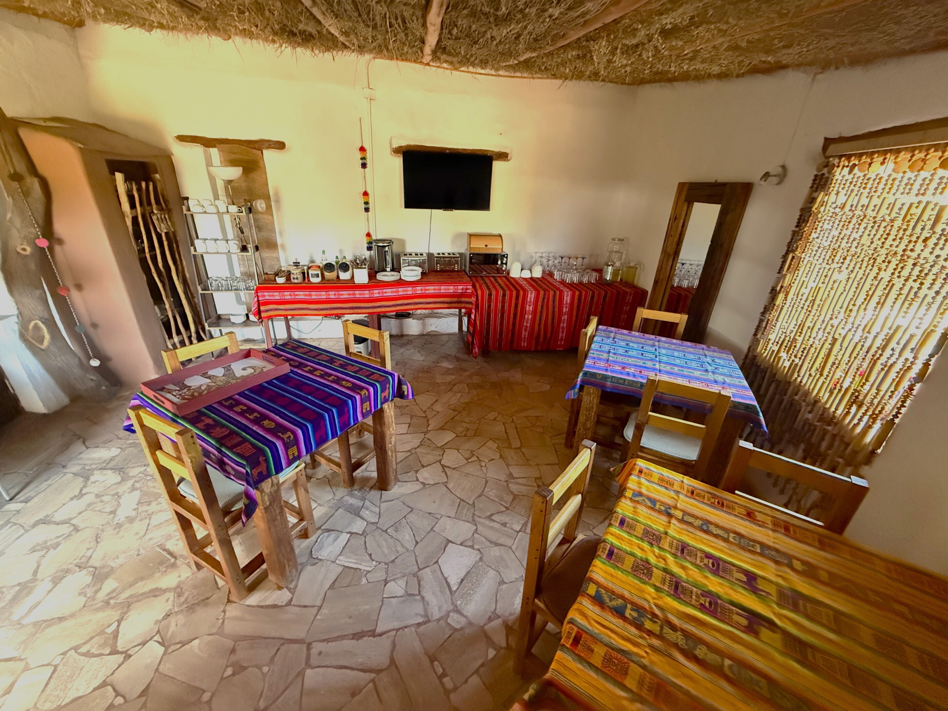 Un comedor con suelo de piedra, mesas de madera con manteles de colores y un televisor montado en la pared.