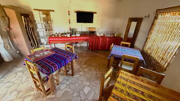 Un comedor con suelo de piedra, mesas de madera con manteles de colores y un televisor montado en la pared.