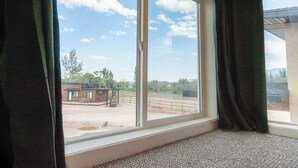 Ein Zimmer mit großem Fenster, Teppichboden und Blick auf einen Außenbereich mit Gebäude und Bäumen.