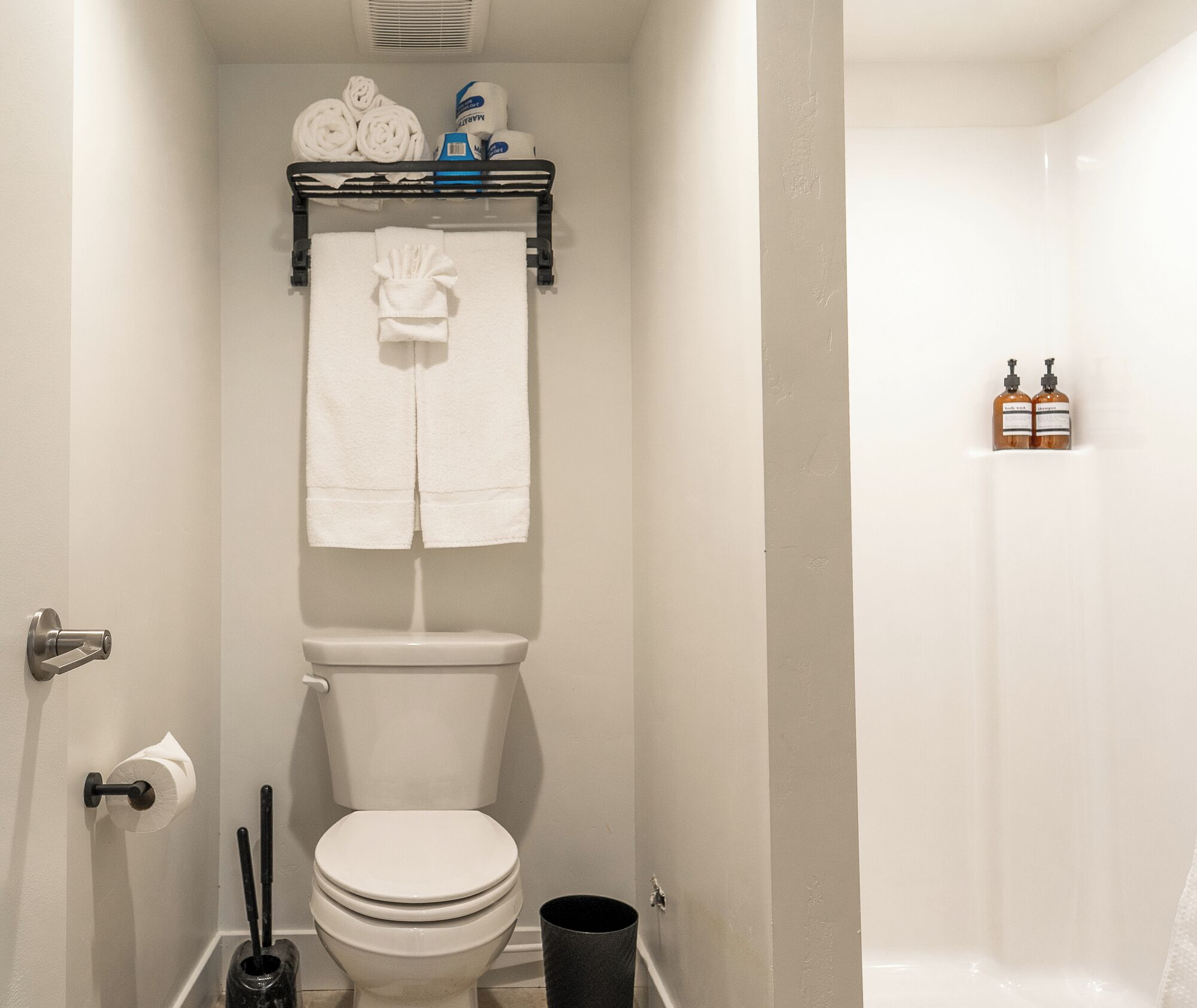 Een badkamer met een toilet, een handdoekrek met handdoeken, een toiletrolhouder, een prullenbak en een zeepdispenser.