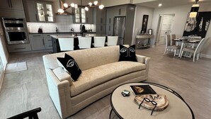 Ein modernes Wohnzimmer mit einem beigefarbenen Sofa, schwarzen Zierkissen, einem runden Couchtisch mit Büchern und Gläsern sowie einem Essbereich mit einer Pendelleuchte.