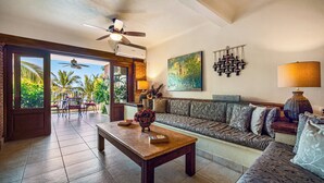 A spacious living area with a sofa, a coffee table, and a ceiling fan. There is a view of a balcony with palm trees and a painting on the wall.
