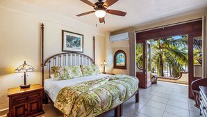 A bedroom with a bed, a ceiling fan, a nightstand, a lamp, a framed picture, and a view of palm trees through a door.