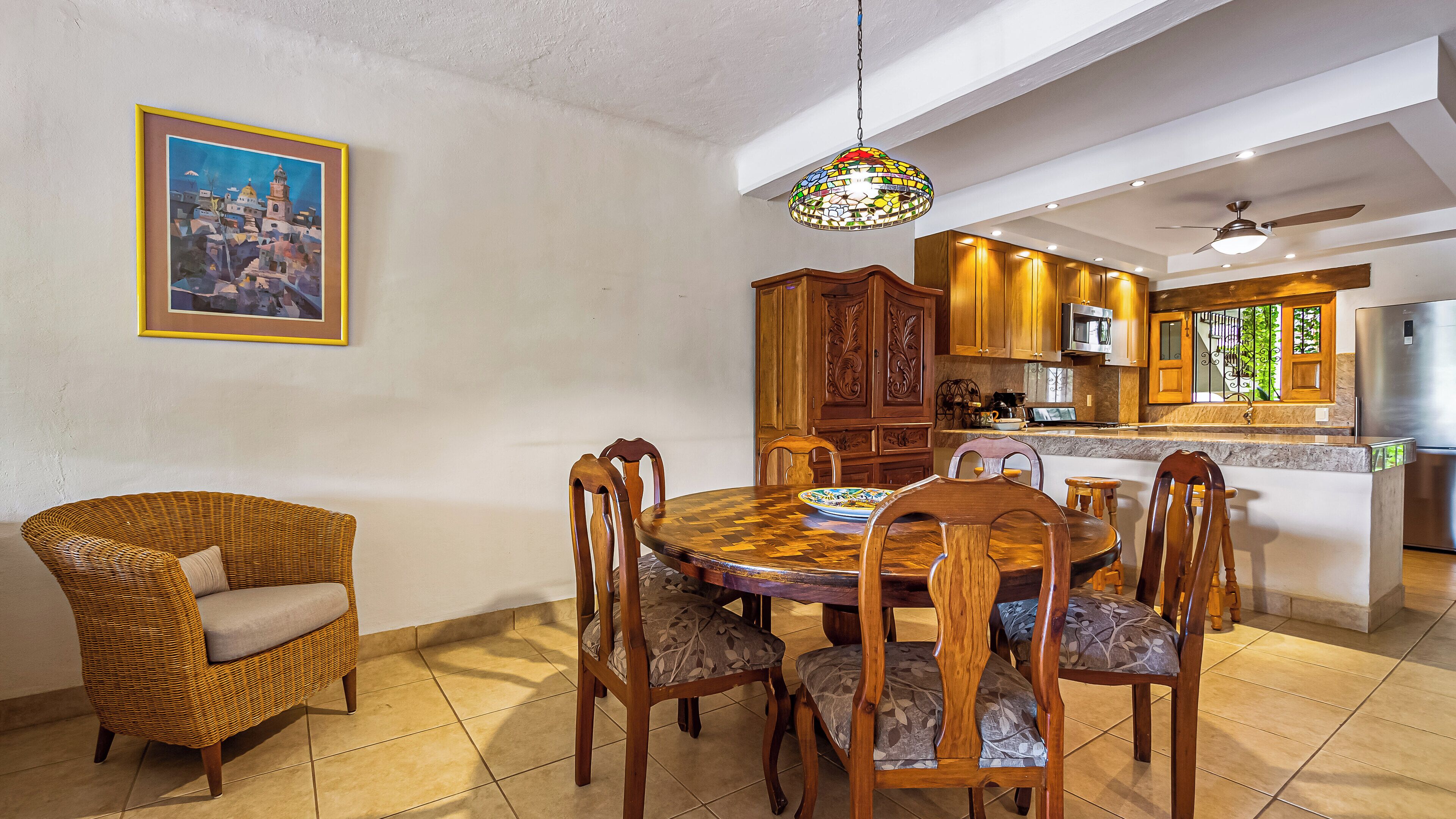 Un coin repas avec une table ronde en bois et des chaises, un fauteuil en osier, un luminaire suspendu en vitrail, et une cuisine en arrière-plan.