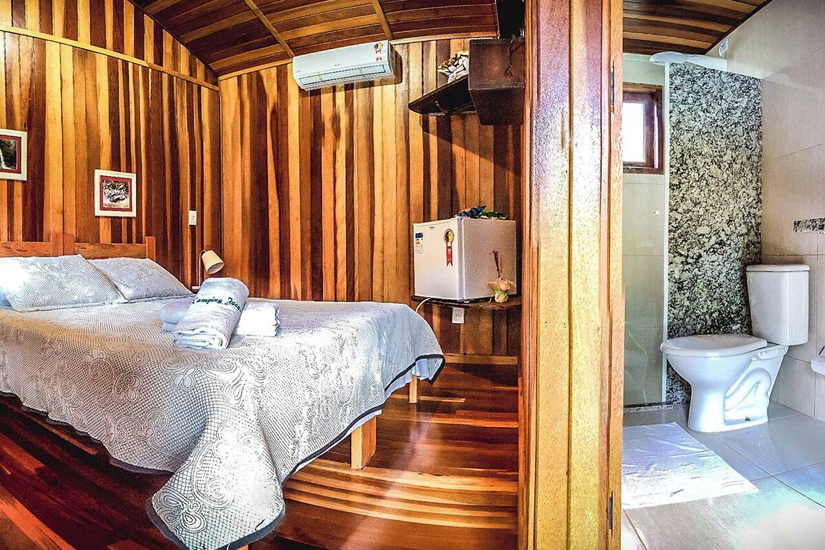 Interior de una cabaña de madera con una cama, un refrigerador y un baño que incluye inodoro y lavamanos.
