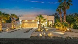 A modern two-story house with a garage, surrounded by palm trees and a well-lit driveway. - Gorgeous Tranquil Desert Retreat (Desert Hot Springs)