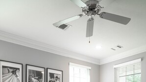 Un dormitorio con una cama, un banco negro, un ventilador de techo y tres cuadros en la pared.