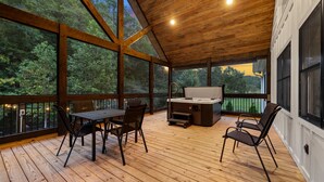 A covered deck with a wooden table and chairs, a hot tub, and large windows offering a view of the surrounding forest.