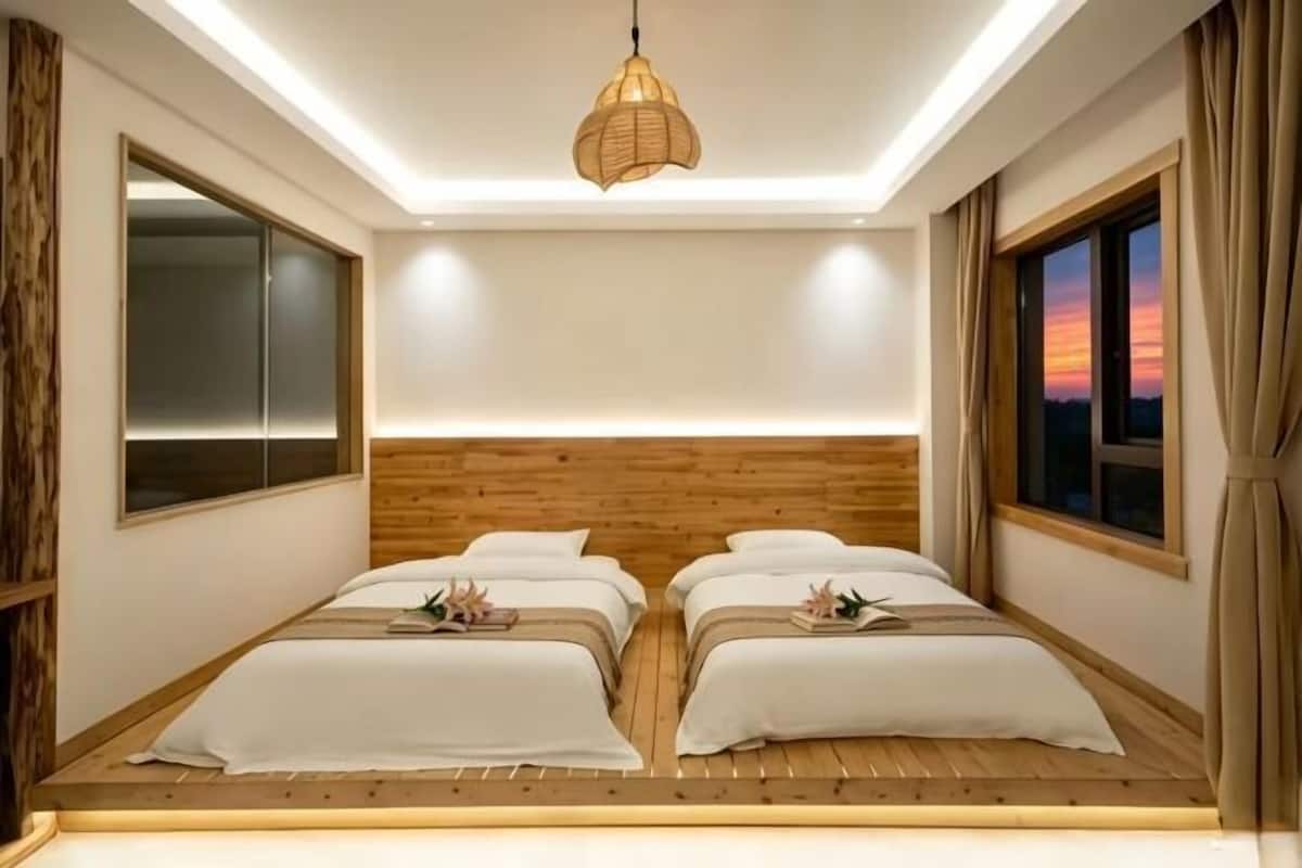 Une chambre avec une tête de lit en bois, deux lits avec du linge de lit blanc, un luminaire suspendu et une fenêtre avec des rideaux.
