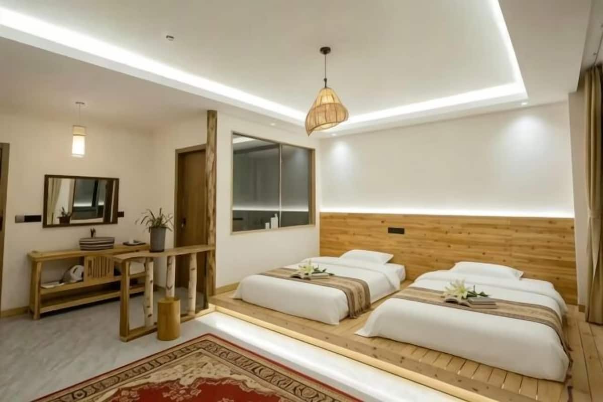 Une chambre d’hôtel moderne avec une tête de lit en bois, un grand miroir, une table de chevet agrémentée d’une plante et un tapis rouge à motifs.