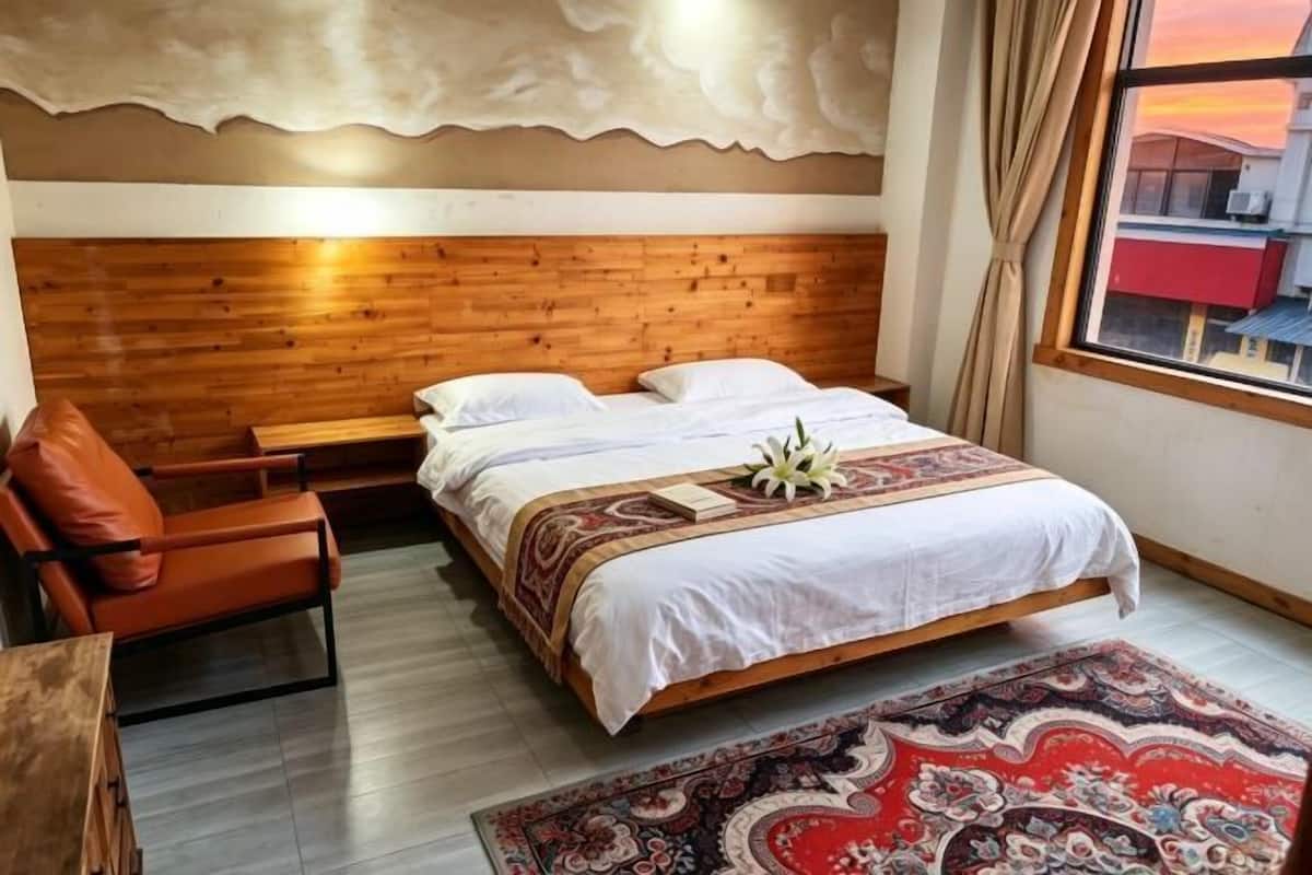 Une chambre avec une tête de lit en bois, un lit recouvert de linge de lit blanc, un tapis rouge à motifs, une table de chevet en bois et une chaise.