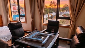 Una habitación con cama, dos sillas, una mesa con un juego de mesa y vistas a un atardecer a través de amplias ventanas.