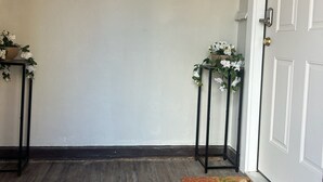 A hallway with a wooden floor, a welcome mat, two small tables with white flowers, and a wall-mounted decorative piece.