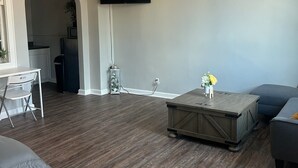 A living room with a ceiling fan, a flat-screen TV mounted on the wall, a dark wood floor, a grey sofa, a coffee table, and a small plant.