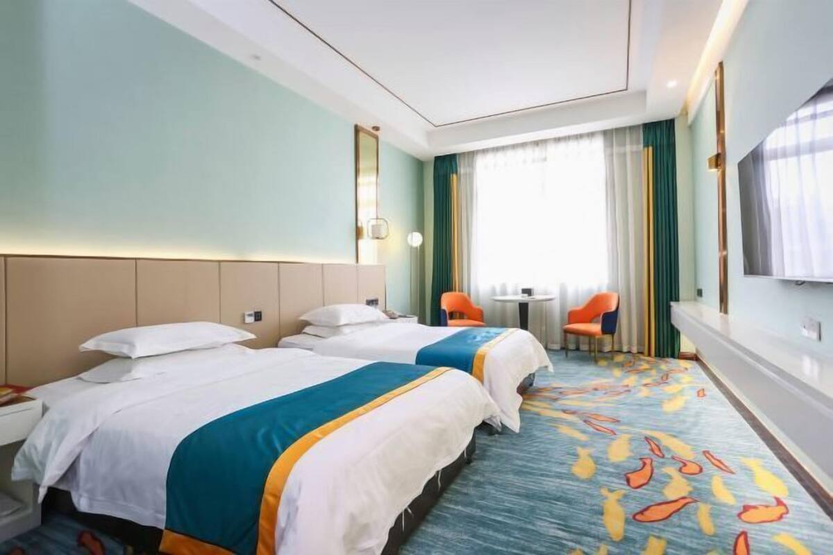 Hotellihuone, jossa on kaksi sänkyä, pieni pöytä, kaksi tuolia, televisio ja ikkuna, jossa on verhot.