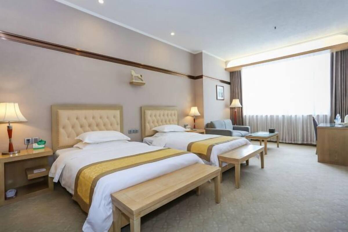 Hotellihuone, jossa on kaksi sänkyä, työpöytä, tuoli, penkki ja suuri ikkuna.