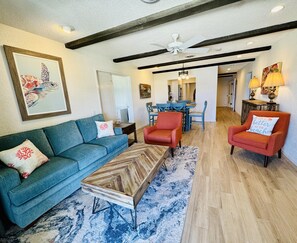 A living room with a blue sofa, a red armchair, and a wooden coffee table. There is a dining area with blue chairs and a chandelier.
