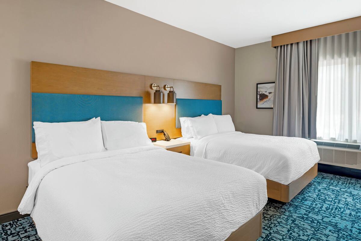 Een hotelkamer met twee bedden, een nachtkastje, een wandlamp en een groot raam met gordijnen.