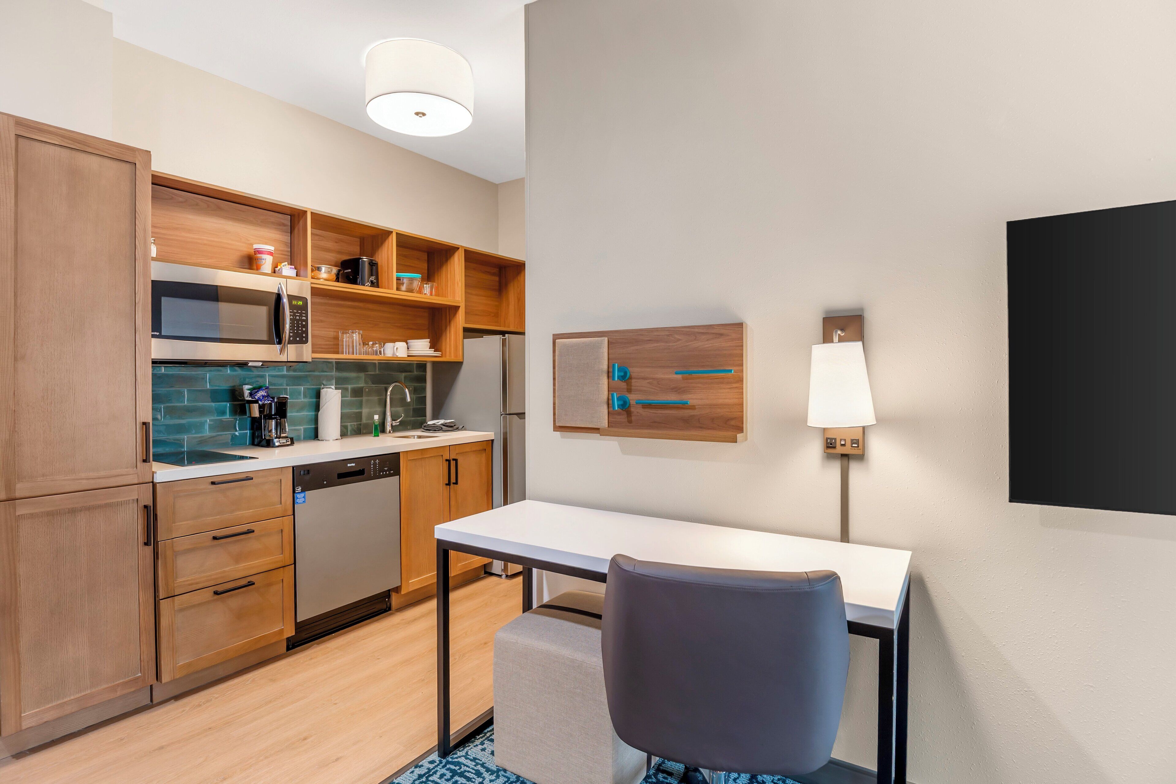Cocina compacta con armarios de madera, microondas, cafetera y lavavajillas. Hay una mesa pequeña con una silla y una lámpara de pared.