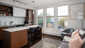 A modern kitchen with a central island, bar stools, and a dining table. There are large windows providing natural light and a view of the outdoors. - Bright Condo in Victoria's Trendy Old Town (Victoria North)