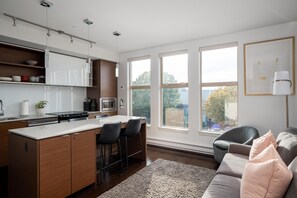 A modern kitchen with a central island, bar stools, and a dining table. There are large windows providing natural light and a view of the outdoors. - Bright Condo in Victoria's Trendy Old Town (Victoria North)