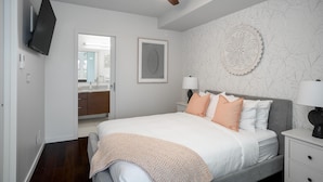 A bedroom with a bed, bedside tables, a ceiling fan, a TV mounted on the wall, and a bathroom visible through an open door. - Bright Condo in Victoria's Trendy Old Town (Victoria North)