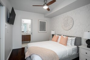 A bedroom with a bed, bedside tables, a ceiling fan, a TV mounted on the wall, and a bathroom visible through an open door. - Bright Condo in Victoria's Trendy Old Town (Victoria North)