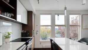 A modern kitchen with a central island, built-in appliances, and large windows offering a city view. - Bright Condo in Victoria's Trendy Old Town (Victoria North)
