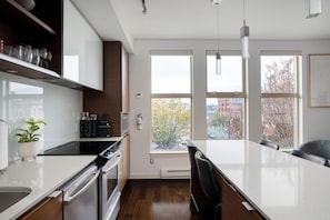 A modern kitchen with a central island, built-in appliances, and large windows offering a city view. - Bright Condo in Victoria's Trendy Old Town (Victoria North)