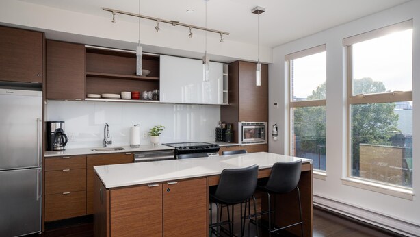 A modern kitchen with a central island, bar stools, and a large window providing a view of the outdoors. - Bright Condo in Victoria's Trendy Old Town (Victoria North)