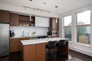 A modern kitchen with a central island, bar stools, and a large window providing a view of the outdoors. - Bright Condo in Victoria's Trendy Old Town (Victoria North)