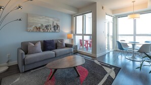 A modern living room with a grey sofa, a coffee table, a cityscape painting, and a dining area with chairs and a table. - Panoramic City Views and Modern Style Condo (Victoria Central British Columbia Provincial Government)