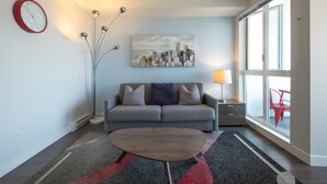 A modern living room with a grey sofa, a coffee table, a cityscape painting, a red clock, and a large window with a view. - Panoramic City Views and Modern Style Condo (Victoria Central British Columbia Provincial Government)