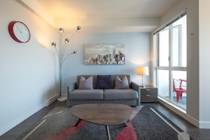 Ein modernes Wohnzimmer mit einem grauen Sofa, einem Couchtisch, einem Stadtbildgemälde, einer roten Uhr und einem großen Fenster mit Blick.