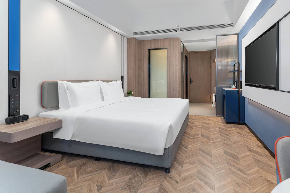 A modern hotel room with a bed, bedside table, TV, and a desk area.