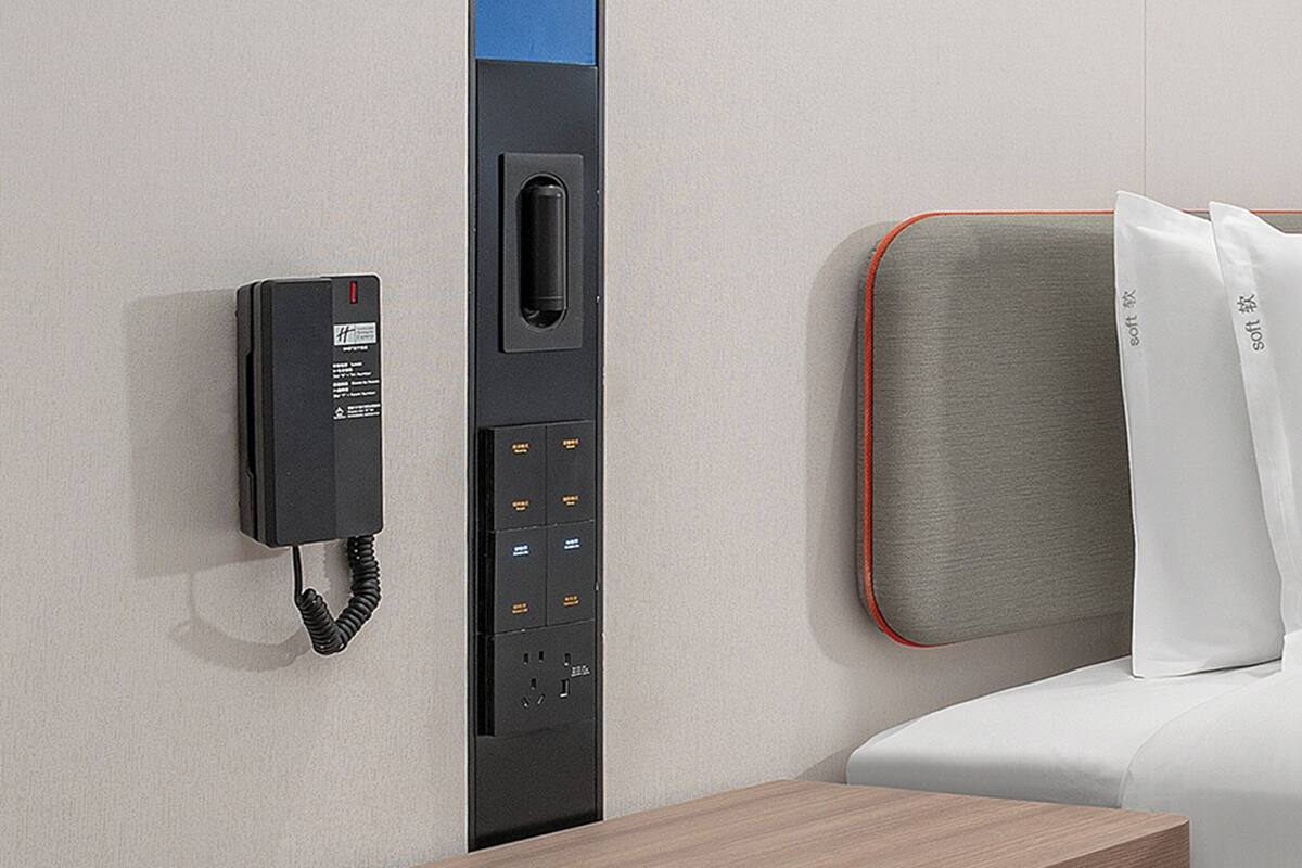 A hotel room with a built-in telephone, a control panel, and a bed with white bedding and a grey headboard.