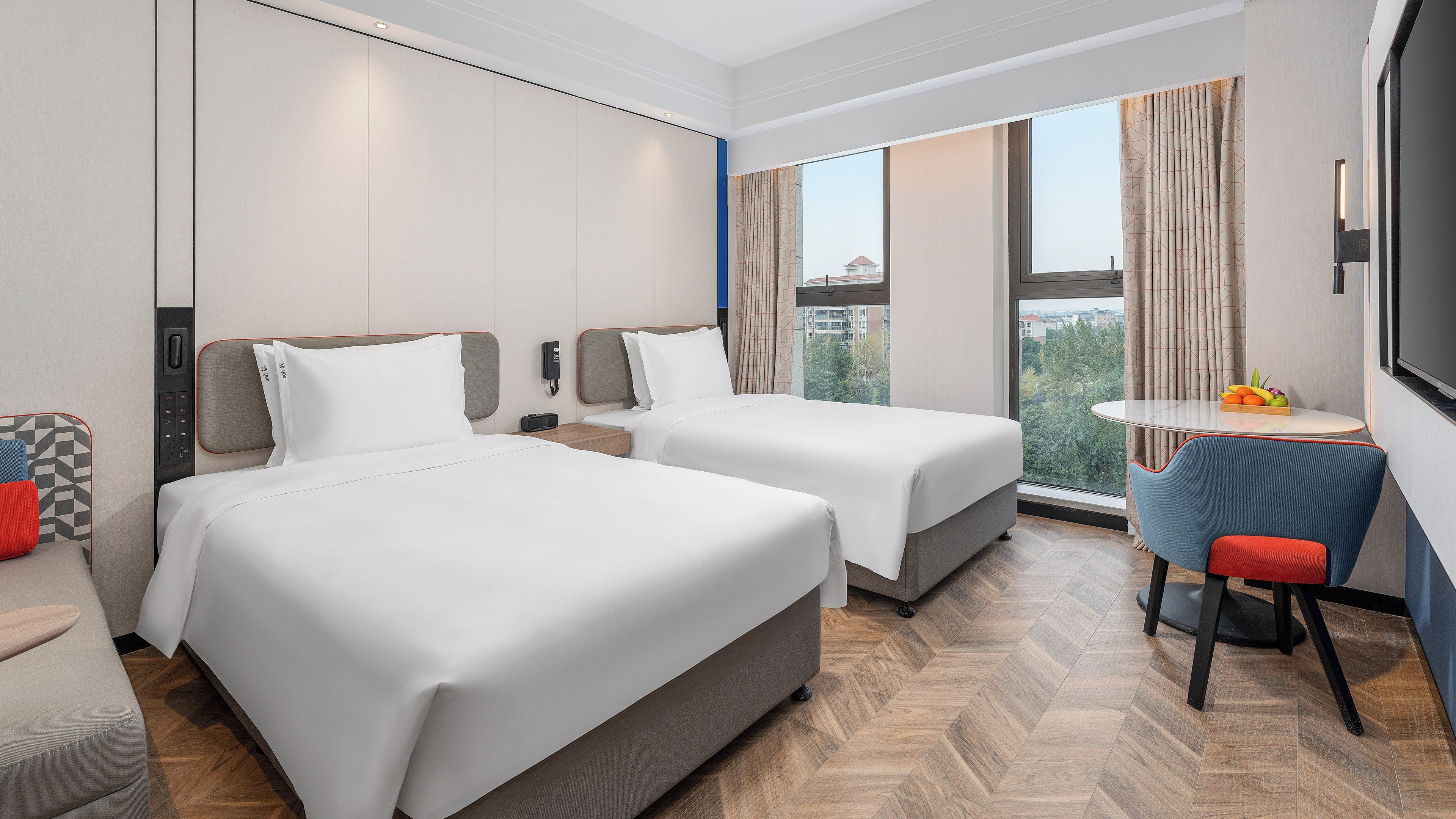 A hotel room with two beds, a desk, a chair, and a large window with a city view.