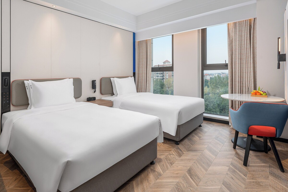 A hotel room with two beds, a desk, a chair, and a large window with a city view.