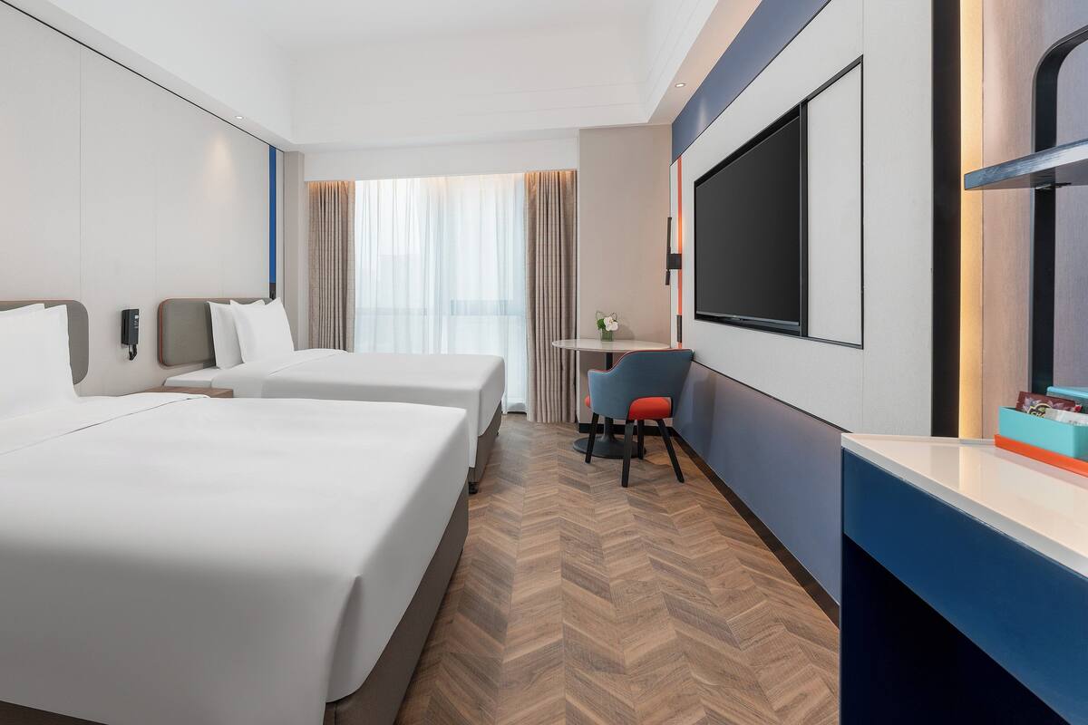 A hotel room with two beds, a desk, a chair, a TV, and a window with curtains.